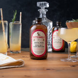 Bittermilk cocktail mixer bottle on a wooden table with cocktails and ingredients.