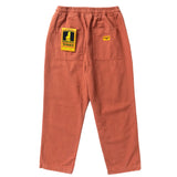 Back of a pair of deep orange straight leg pants with a drawstring waist and two back pockets. The left pocket has a yellow and black cardboard tag covering it with a cartoon of a waiter and the text [Service Works]. The right pocket has a small orange patch sewn onto it with the Service Works logo. 