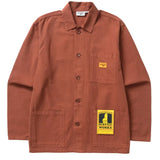 Front of a canvas chore style jacket in a deep orange color with two hand pockets and a chest pocket on the wearer's left side. The chest pocket has a small orange fabric patch on it with the Service Works logo. Over the wearer's left hand pocket is a yellow and black cardboard tag with a cartoon waiter and the text [Service Works] on it. 