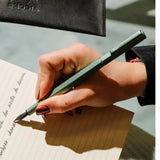 A close-up of a hand writing in a lined notebook with a metallic green pen, with a black Riotta pouch partially visible at the top.