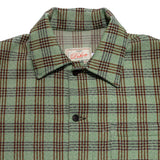 Close up of collar of a button up shirt in green and brown plaid. There are two small dark buttons visible in the center, as well as a white fabric tag at the back of the neck with [1920 Dehen] on it, and a chest pocket on the wearer's left side. 