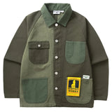 Front of a canvas chore-style jacket with silver buttons color blocked in various shades of green. There are two chest pockets, one with a silver button, and two hand pockets. On the right hand pocket there is a yellow and black cardboard tag with a cartoon waiter and the text [Service Works] and above that same pocket is a small white fabric patch with the Service Works logo. 