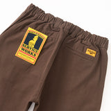 Back of pair brown cotton canvas pants with two welt back pockets. Over the left pocket is a yellow and black cardboard tag with a cartoon waiter and the text [Service Works] and above the right pocket is a small orange fabric patch with the Service Works logo